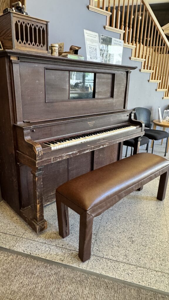 Photo of an upright piano in front of a gray-painted wall and a staircase.